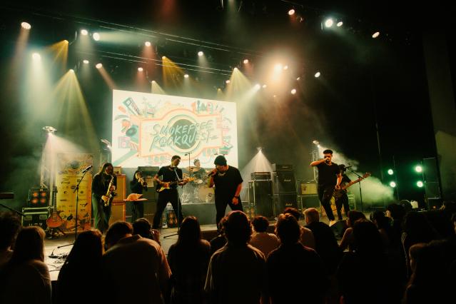 A band playing on a stage with the SmokefreRockquest logo projected behind them.